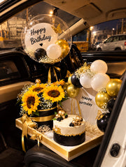 Luxury sunflower birthday arrangement with black and gold ribbons, white balloons, and decorated cake.