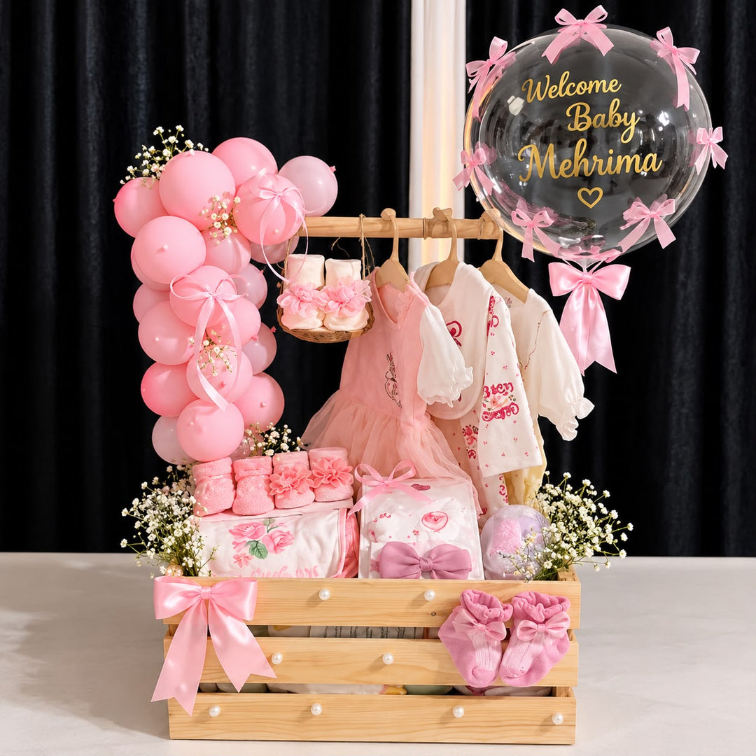 Baby girl gift hamper in a wooden crate with pink outfits, booties, flowers, and a clear balloon reading “Welcome Baby Mehrima.”