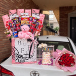 Load image into Gallery viewer, Pink butterfly chocolate and snack hamper with a teddy bear, displayed beside a boxed celebration cake and pink-wrapped red roses.