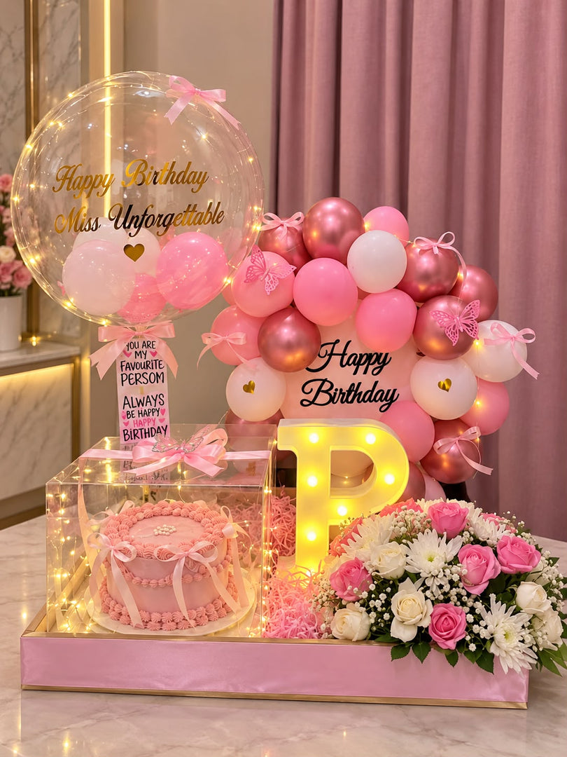 Pink birthday hamper with a cake in a clear box, pink and white flowers, birthday balloons, fairy lights and an illuminated letter P.