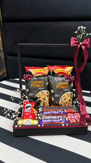Black gift hamper with caramel peanut popcorn, Pringles, Toblerone, Dairy Milk, KitKat and Ferrero Rocher, finished with burgundy bows and white flowers.