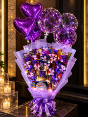 Lavender-wrapped chocolate and snack bouquet with KitKat, Snickers, Danisa cookies, purple bows, heart balloons and illuminated confetti balloons.