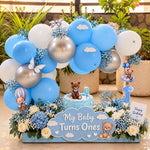 Load image into Gallery viewer, Blue first birthday arrangement with a teddy bear cake, number-one candle, blue and silver balloon arch, ivory roses and a cloud-shaped sign.