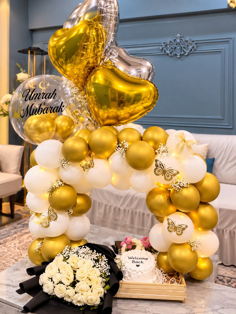 Gold and white Umrah Mubarak balloon arrangement with heart-shaped foil balloons, a Welcome Back cake and an ivory flower bouquet on a wooden tray.