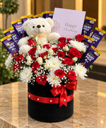 Load image into Gallery viewer, Red roses and white chrysanthemums arranged with a white teddy bear and Dairy Milk chocolates in a black gift box with a red ribbon.