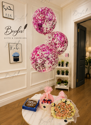Three round pink and white patterned balloons tied to a table in a decorated indoor room, alongside a chocolate cake, a wrapped gift basket, and a bouquet of flowers. The scene has visible 'Beegles!' watermarks and framed wall art in the background.
