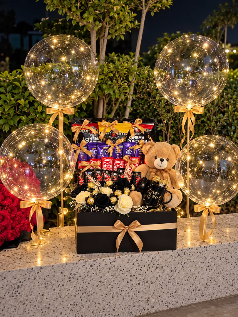 Black and gold gift hamper with chocolates, popcorn, a teddy bear, Love You mug and cream and black roses, surrounded by four illuminated balloons.