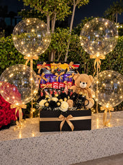 Black and gold gift hamper with chocolates, popcorn, a teddy bear, Love You mug and cream and black roses, surrounded by four illuminated balloons.