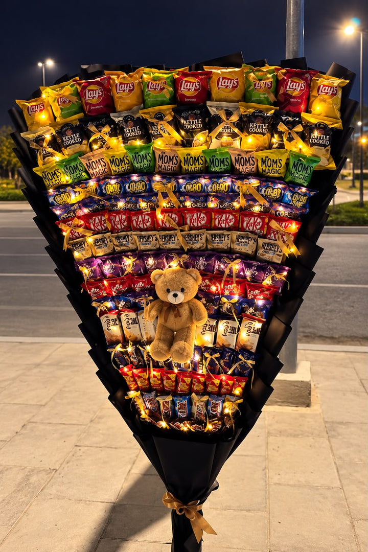 Giant black-wrapped snack bouquet with Lay’s crisps, biscuits, assorted chocolate bars, a brown teddy bear, gold bows and warm fairy lights.