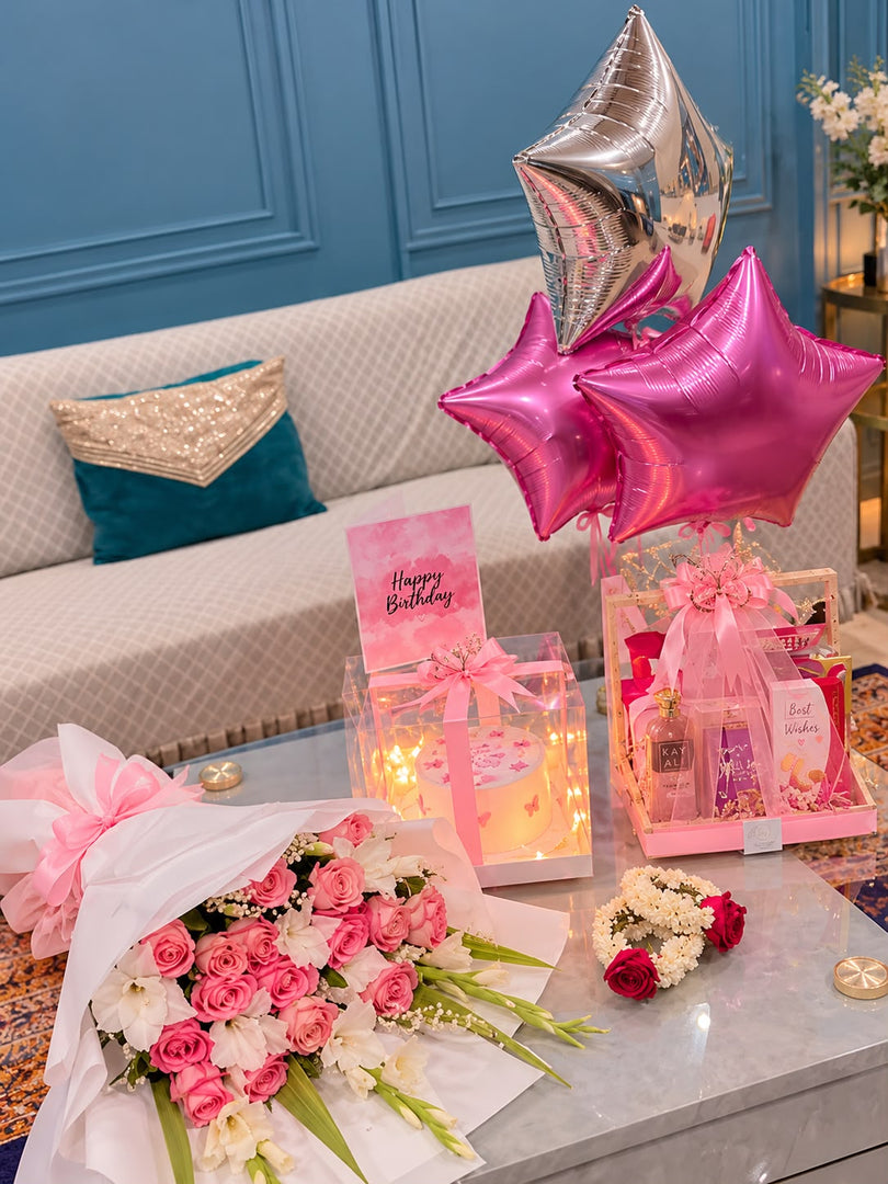Pink birthday gift set with a rose and white flower bouquet, cake in a lighted clear box, perfume basket, floral garlands and pink and silver star balloons.