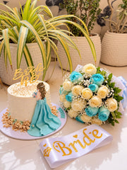 Ivory bridal shower cake with a turquoise gown decoration and gold Bride to Be topper beside an ivory and turquoise rose bouquet and Bride sash.

