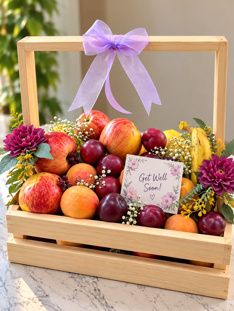 Fresh Wishes get-well-soon fruit basket with red apples, bananas, plums, citrus fruits, purple flowers, lavender bow, and wooden crate.