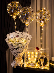 Black tuxedo cake, Ferrero Rocher chocolates and illuminated W in a clear gift box beside white flowers and three lighted gold confetti balloons.