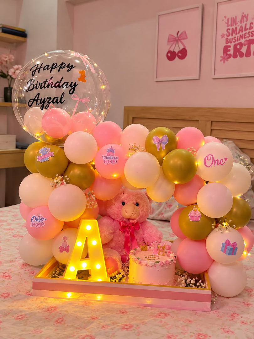Pink first birthday hamper with a teddy bear, number one cake, pink and gold balloons, a birthday message balloon and an illuminated letter A.