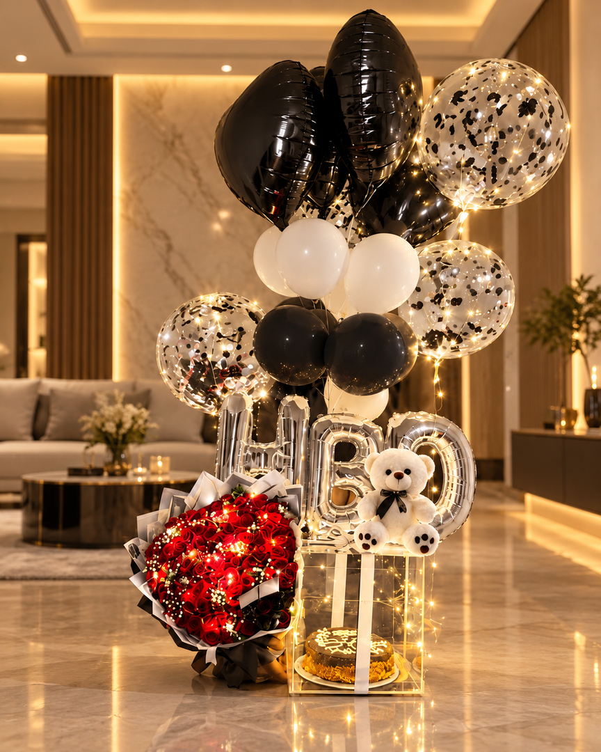 Birthday gift display with red roses, a cake in a clear box, white teddy bear, silver HBD letters and black and white balloons with fairy lights.
