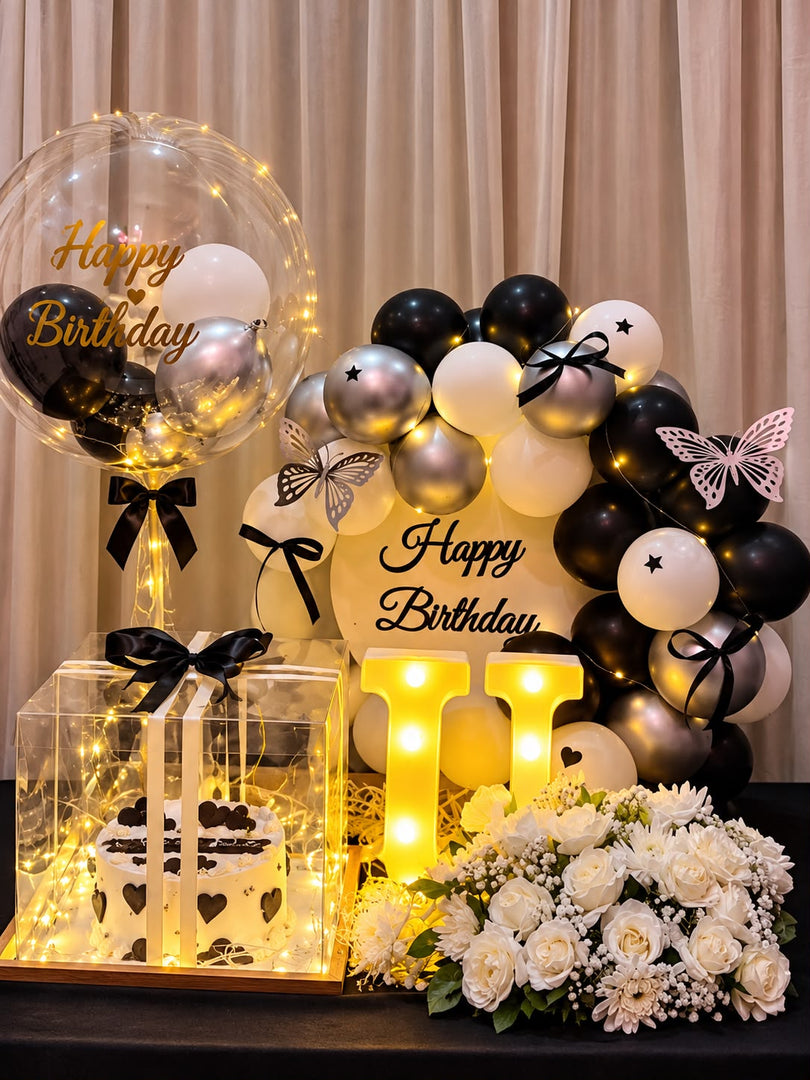 Black and white birthday hamper with a heart-decorated cake in a clear box, white roses, silver balloons, fairy lights and an illuminated U.