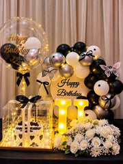 Black and white birthday hamper with a heart-decorated cake in a clear box, white roses, silver balloons, fairy lights and an illuminated U.
