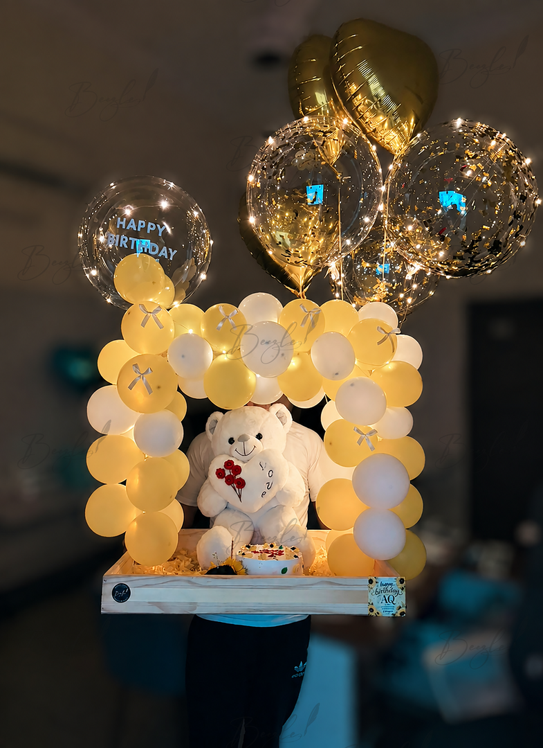 Premium birthday surprise featuring a glowing balloon arch, teddy bear, birthday cake, confetti balloons, and elegant wooden display tray.