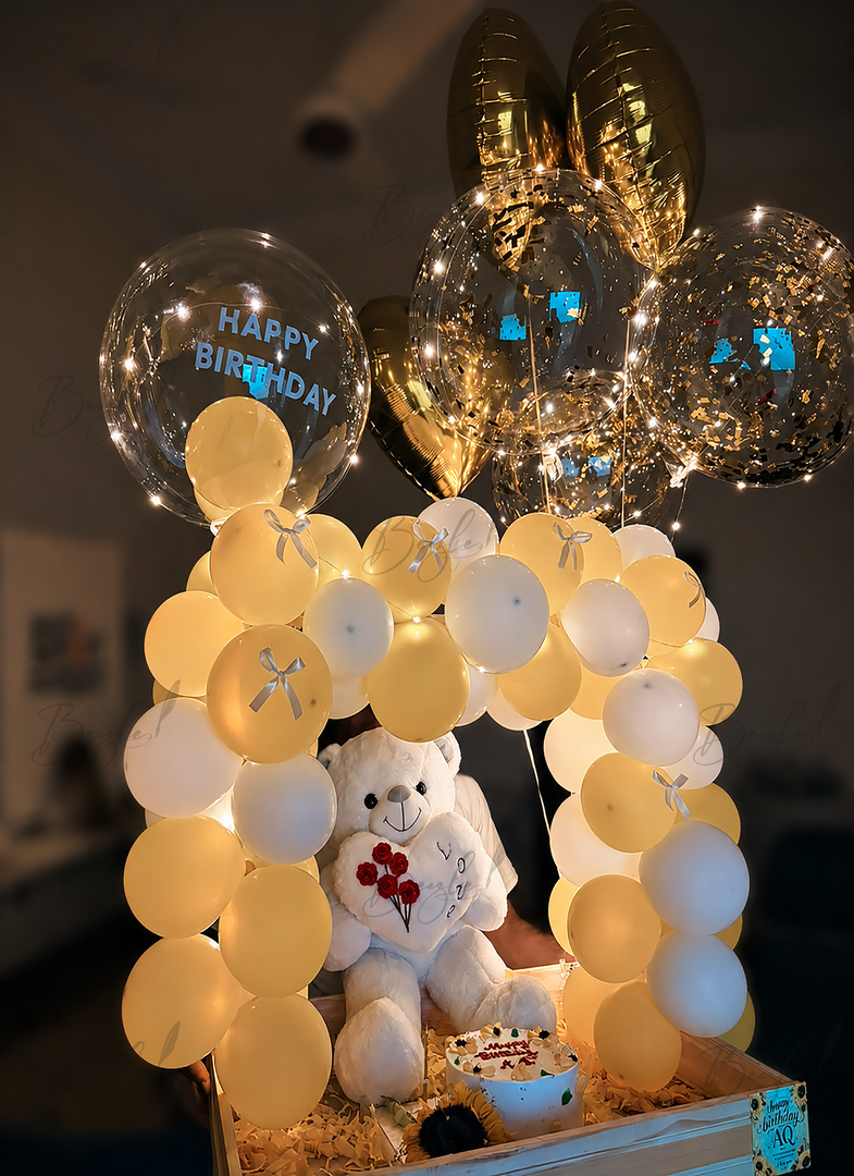 Luxury birthday gift setup with LED balloon arch, white teddy bear, birthday cake, gold chrome balloons, and illuminated balloon decorations.