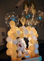 Luxury birthday gift setup with LED balloon arch, white teddy bear, birthday cake, gold chrome balloons, and illuminated balloon decorations.