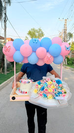 Load image into Gallery viewer, Person holding a tray with balloons, birthday cakes, and flower bouquet on a street.