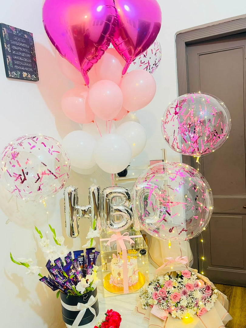 Decorative arrangement of balloons, flower bouquet, birthday cake and a 'HBD' balloon with a chocolate gift box on side.