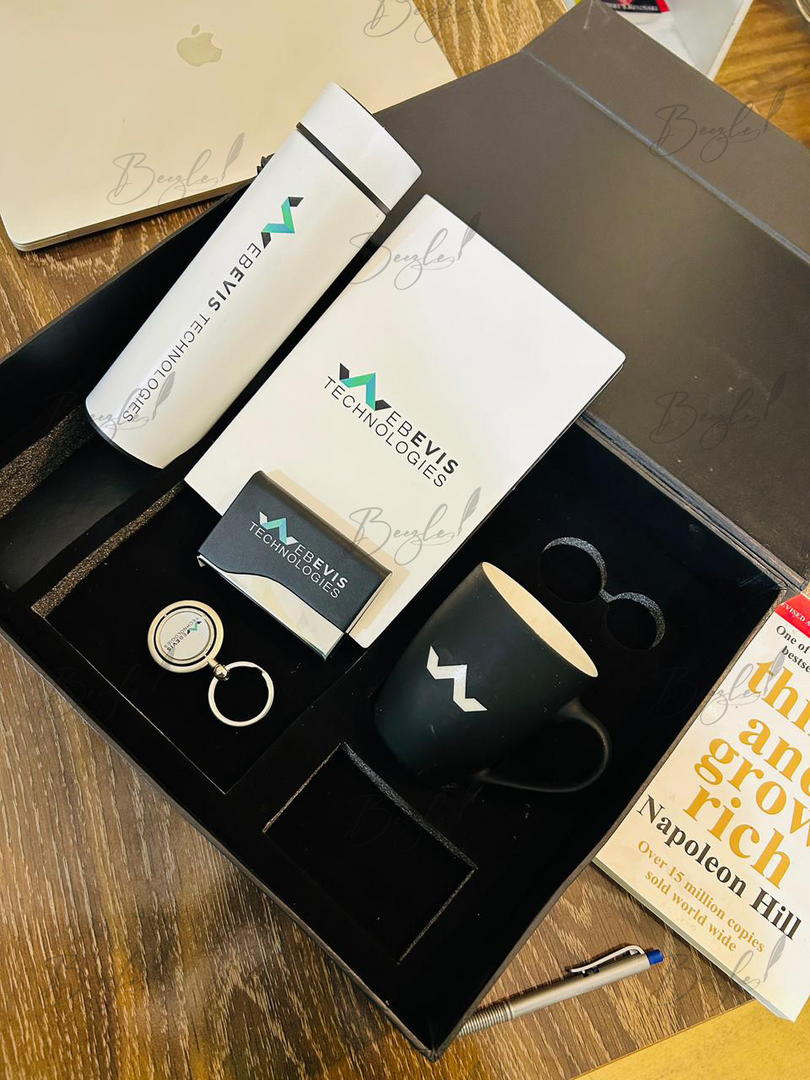 A branded gift set from Webevis Technologies displayed in a black presentation box on a wooden table. The set includes a black ceramic mug with a white logo, a white branded box, a black business card, a round keychain, and a pen.