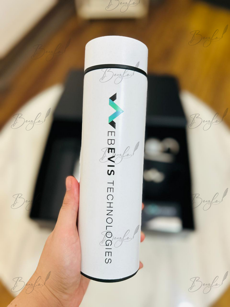 A hand holding a white cylindrical bottle with black text reading 'WEBEVIS TECHNOLOGIES' and a small teal and black logo, against a blurred indoor background with a wooden floor and white table surface.