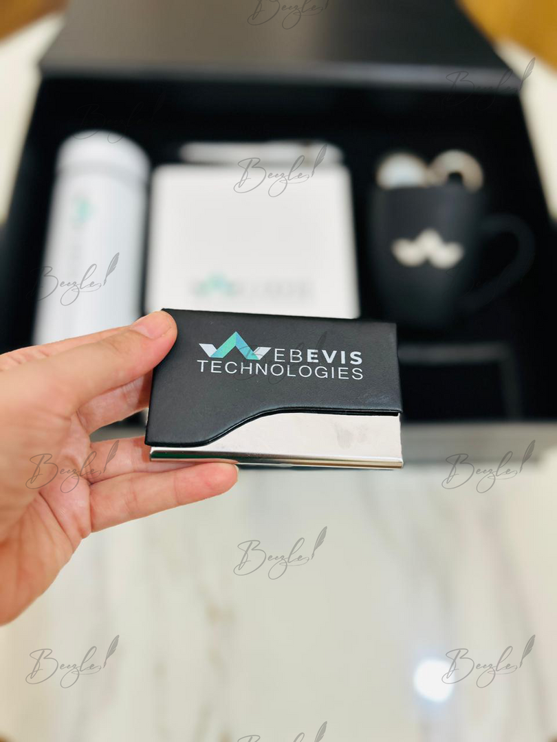 A hand holds a black and silver metal business card for 'WEbevis Technologies' in front of a blurred gift set containing a white bottle, a white box, and a black mug.