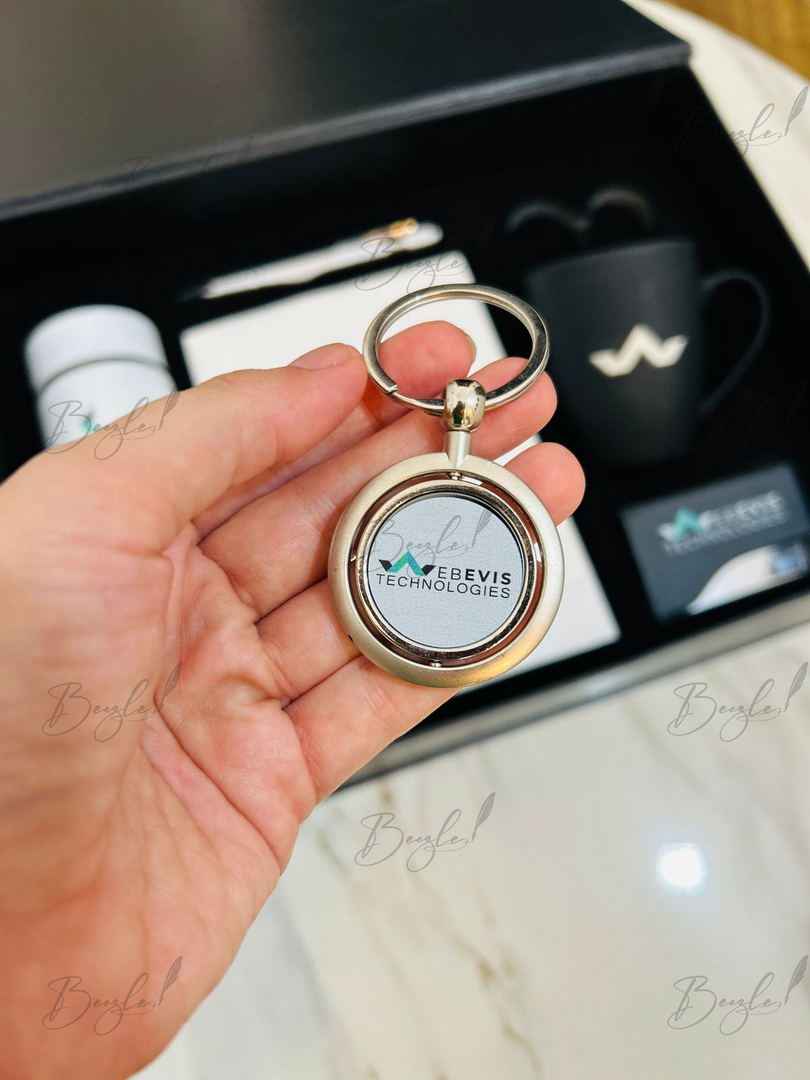 A hand holds a round silver-toned keychain with a logo insert reading 'WEBEVIS TECHNOLOGIES' in front of a gift box containing a black mug, white bottle, and card.