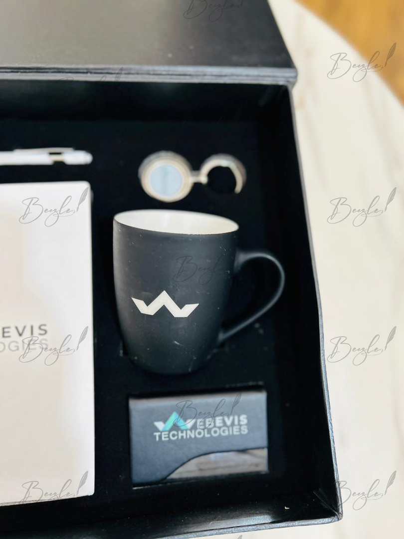 A black gift set box containing a matte black ceramic mug with a white logo, a white branded card, a small round item, and a black card labeled 'Beavis Technologies'.