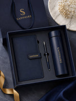 Load image into Gallery viewer, A Sapphire-branded gift set in a dark navy box, including a matching notebook, pen, and insulated tumbler, with a branded shopping bag and decorative ribbon and flowers in the background.