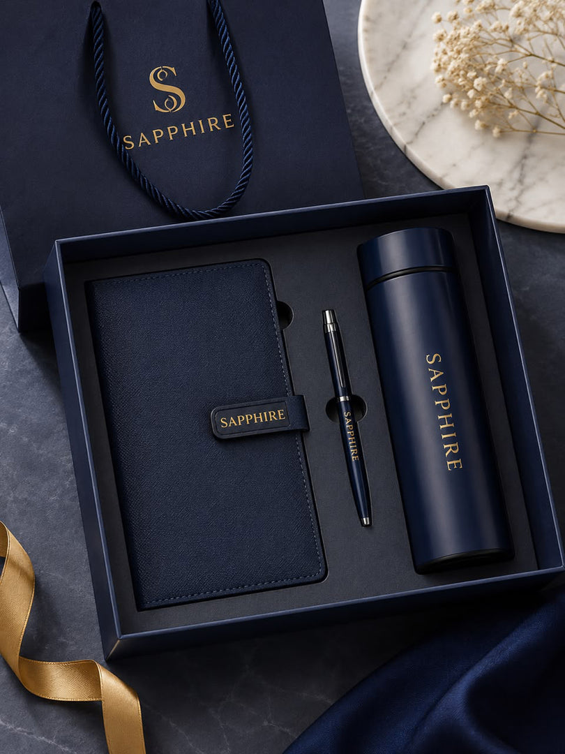 A Sapphire-branded gift set in a dark navy box, including a matching notebook, pen, and insulated tumbler, with a branded shopping bag and decorative ribbon and flowers in the background.