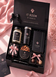 A luxury gift set from IT ROOM BY WEBEVIS, presented in an open black box with pink satin ribbons. The set includes a candle, a tin, a notebook, chocolates, a pen, and a small gift box, all branded with the IT ROOM logo.