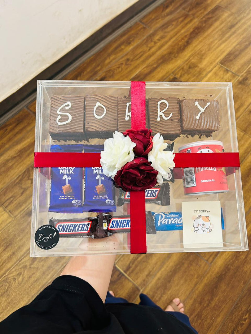 Sorry treat box with chocolate brownies, assorted chocolates, snacks, flowers, and an apology card in a transparent gift box