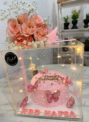 A pink birthday cake displayed inside a clear acrylic gift box with built-in warm fairy lights, decorated with pink butterflies, white pearls, and a bouquet of peach and white flowers on top. The box has 'HBD HAFSA' written on the base, and a black circular logo sticker is visible on the left side.
