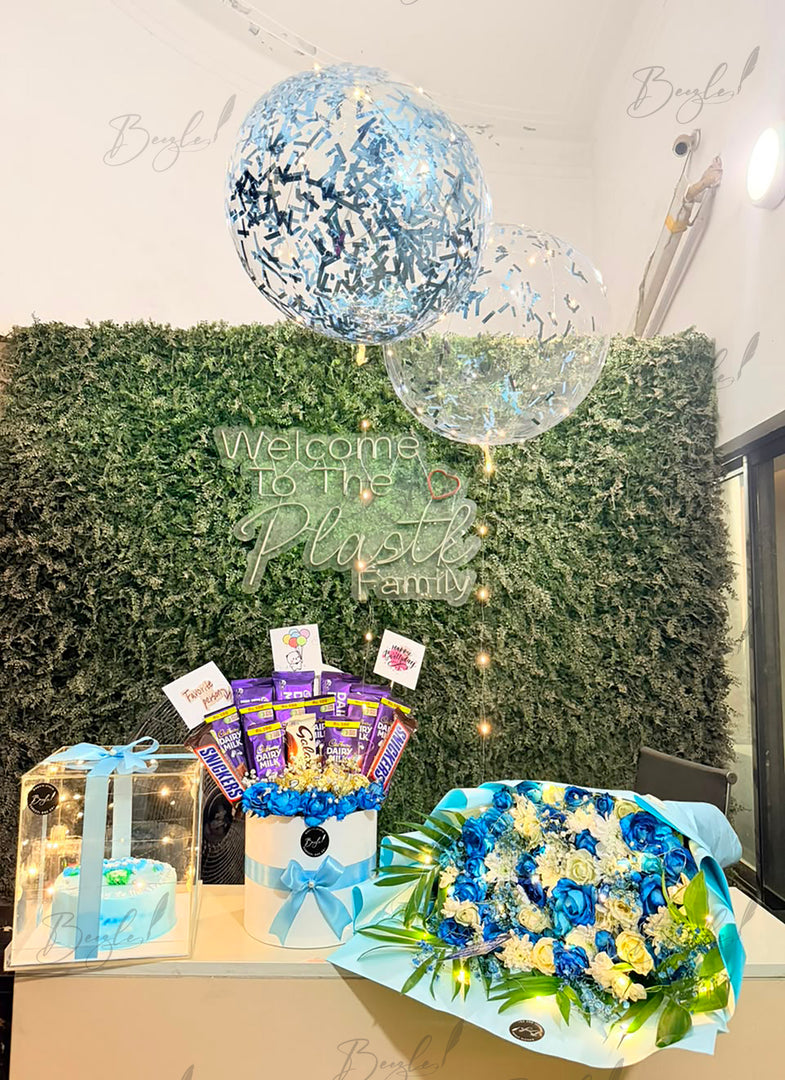 A gift arrangement displayed indoors against a green hedge wall backdrop, featuring a large blue and silver confetti balloon, a clear balloon, a wrapped cake in a clear box with a blue ribbon, a chocolate bucket bouquet with purple candy sticks, and a blue and white flower arrangement.
