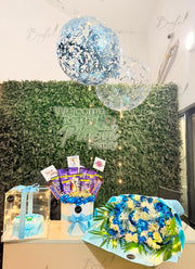 A gift arrangement displayed indoors against a green hedge wall backdrop, featuring a large blue and silver confetti balloon, a clear balloon, a wrapped cake in a clear box with a blue ribbon, a chocolate bucket bouquet with purple candy sticks, and a blue and white flower arrangement.