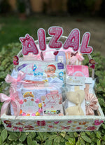 Load image into Gallery viewer, A baby gift basket labeled 'AIZAL' filled with baby items including diapers, wipes, a footie sock, and a small plush toy, arranged in a white basket with pink bows and decorations