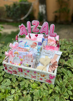 Load image into Gallery viewer, A baby shower gift basket filled with baby essentials, decorated with pink polka-dot lettering that reads 'AIZAL', and placed outdoors on green variegated foliage.