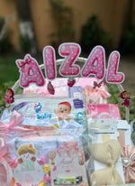 Load image into Gallery viewer, A baby gift basket with a large pink and white polka-dot 'AIZAL' sign, surrounded by baby items including a blue and white baby outfit, a pink and white card, and a beige bow, set against a blurred outdoor background.