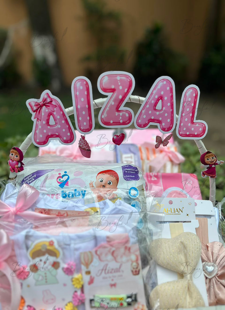 A baby gift basket with a large pink and white polka-dot 'AIZAL' sign, surrounded by baby items including a blue and white baby outfit, a pink and white card, and a beige bow, set against a blurred outdoor background.