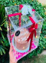 Load image into Gallery viewer, A clear acrylic cake box with built-in fairy lights, red ribbon, and white roses, holding a chocolate birthday cake, held by a hand in front of a green garden background.