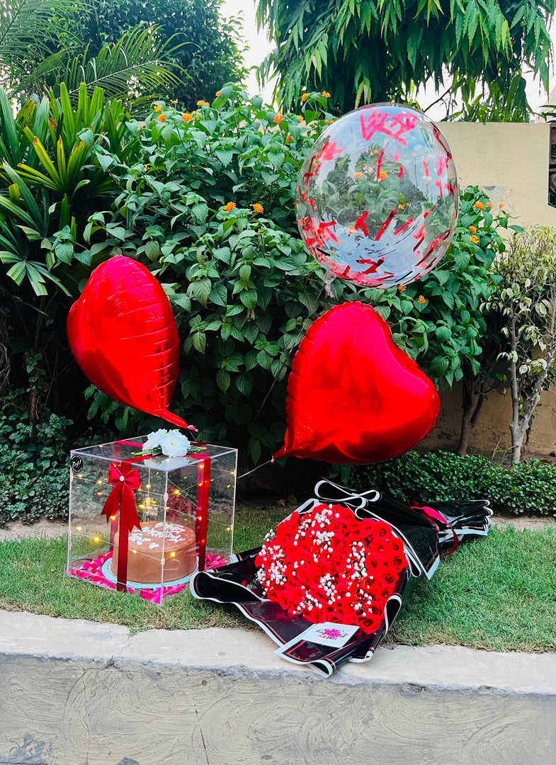 A Birthday gift arrangement featuring two red heart-shaped foil balloons, a clear balloon with red lettering, a decorated red and white floral fabric bag, and a small cake in a clear box, all set on a lawn in front of green bushes.