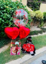Load image into Gallery viewer, A Birthday gift set arranged outdoors on a grassy lawn next to a garden bed. The set includes a large clear balloon filled with red hearts, two red heart-shaped foil balloons, a small wrapped gift box, a black and red patterned fabric item, and a red floral arrangement.