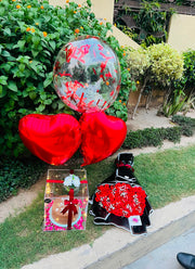 A Birthday gift set arranged outdoors on a grassy lawn next to a garden bed. The set includes a large clear balloon filled with red hearts, two red heart-shaped foil balloons, a small wrapped gift box, a black and red patterned fabric item, and a red floral arrangement.
