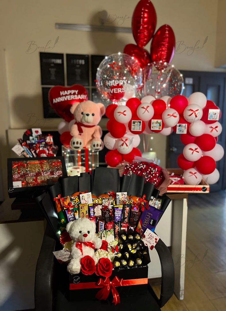 Premium anniversary gift arrangement featuring Ferrero Rocher chocolates, snack bouquet, teddy bear, fresh roses, and romantic balloon decoration