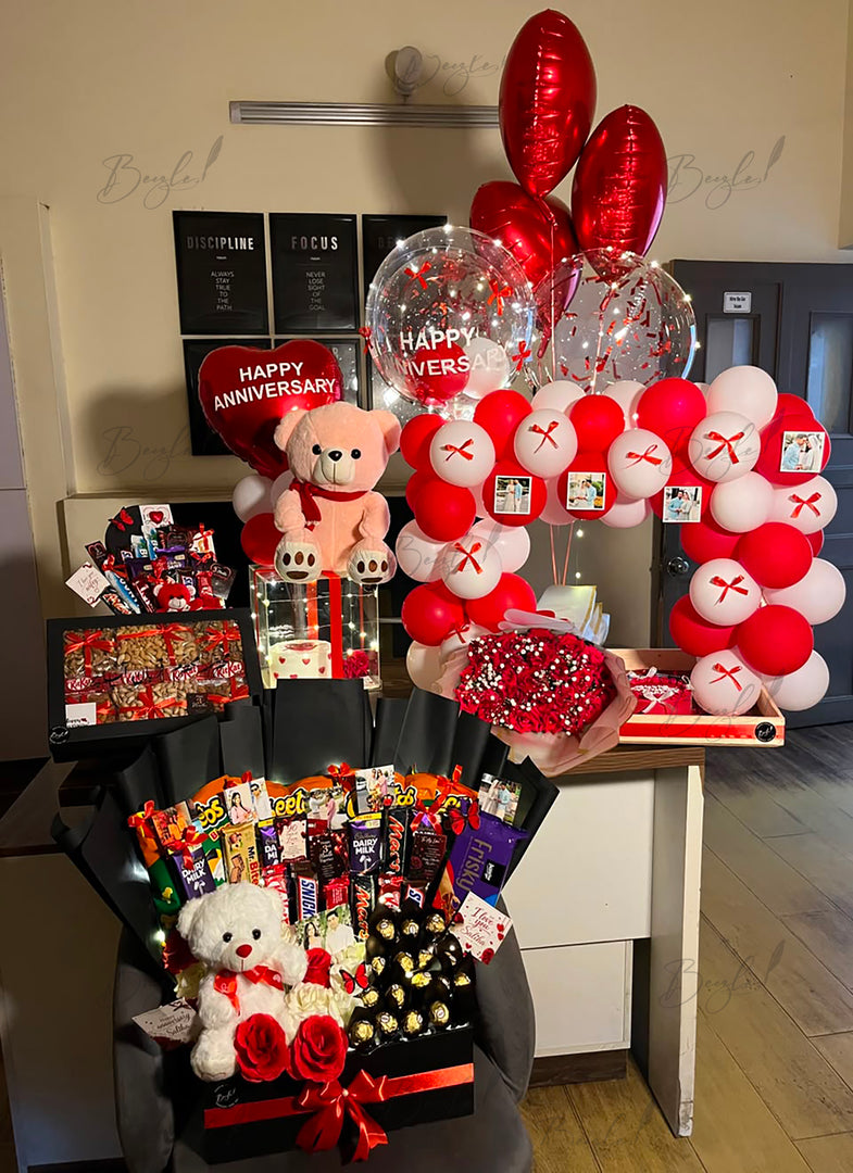 Crimson Promise luxury anniversary gift setup with chocolate gift box, teddy bear, red rose bouquet, personalized balloons, and balloon arch