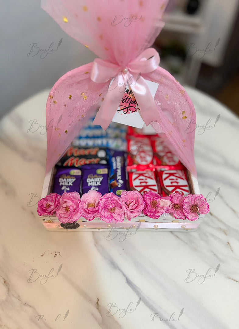 Premium chocolate gift basket with assorted chocolates, pink floral decoration, mesh wrap, and satin ribbon for birthdays and celebrations