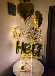 Luxury birthday balloon box with LED lights, transparent acrylic gift box, birthday cake, gold heart balloons, and illuminated confetti balloons.
